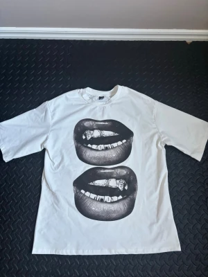 Vit t-shirt med grafiskt tryck - Vit t-shirt med två stora grafiska tryck av läppar i svart på framsidan. Klassisk rund hals och korta ärmar. Tillverkad i mjuk bomull som känns skön mot huden. Perfekt för dig som vill sticka ut med en unik och edgy stil.