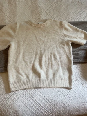Krämvit stickad tröja kort ärm - Mysig krämvit stickad tröja med korta ärmar och rund hals. Tröjan har ribbade muddar vid ärmslut och nederkant, vilket ger en snygg och enkel look. Perfekt att matcha med jeans eller kjol för en clean stil.