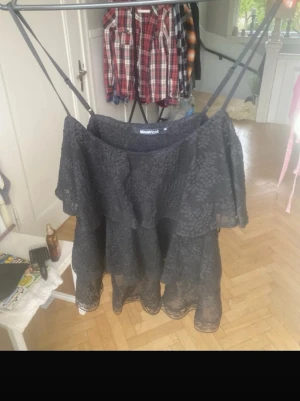 Svart volang linne - Säljer en svart kortklänning med tunna axelband och volangdetaljer. Klänningen är i spetsmaterial och har en luftig, omlottliknande design som ger en romantisk känsla. Perfekt för sommarens alla tillfällen.