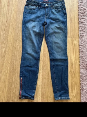Mörkblå skinny jeans med dragkedja Tommy Hilfiger  - Snygga mörkblå skinny jeans Tommy Hilfiger med klassisk femficksmodell och dragkedjedetalj i rött och guld vid benslutet. Jeansen har en lätt tvättad look och smal passform som sitter tajt hela vägen ner. Perfekta till sneakers eller boots. Storleksmärkta 6 och jag som har 36 som vanlig klädstorlek upplever dem som rymliga. 