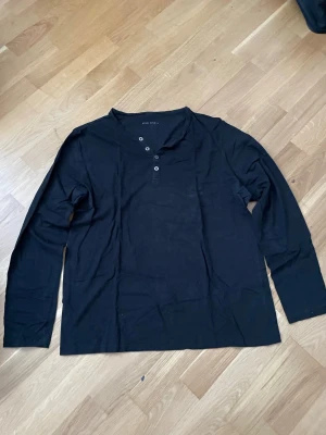 Svart långärmad herrtröja i bomull ,XL - Stilren svart långärmad henleytröja med tre knappar vid halsen. Tröjan har en rak passform och är tillverkad i ett mjukt och bekvämt material, perfekt för en avslappnad look.