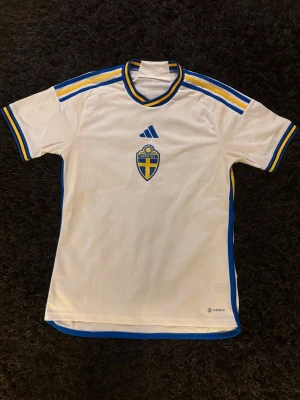 Svensk landslagströja Adidas vit - Vit matchtröja från Adidas med Sveriges landslagsemblem på bröstet. Tröjan har blå och gula detaljer på ärmar och krage samt Adidas-logga framtill. Tillverkad i lätt och ventilerande material, perfekt för fotboll eller träning.