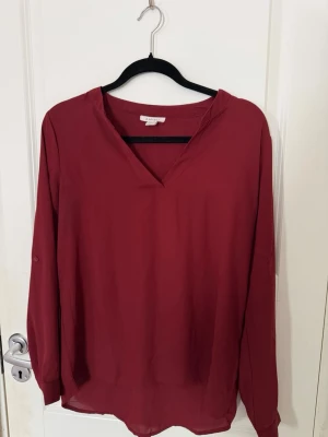 Vinröd blus från Amisu - Stilren vinröd blus från Amisu med v-ringad hals och långa ärmar. Tillverkad i ett lätt och luftigt material som ger en snygg passform. Perfekt att matcha med jeans eller kjol för en trendig look.