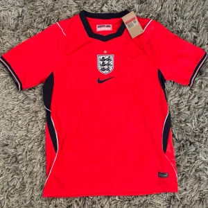 Plick:🏴󠁧󠁢󠁥󠁮󠁧󠁿 England Landslagströja  - Säljer en riktigt snygg röd England-landslagströja från Nike! Riktigt stilren design med mörkblå och vita detaljer, det klassiska Three Lions-emblemet på bröstet och den ikoniska "Happy and Glorious"-detaljen i nacken. 🦁✨ Klockren till både fotbollsmatcher, träning och som streetwear-outfit! 