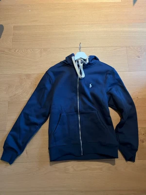 Mörkblå hoodie med dragkedja - Snygg mörkblå hoodie med dragkedja och vita snören i huvan. Klassisk design med fickor framtill och en liten broderad logga på bröstet. Perfekt för en chill och avslappnad stil. Det är st M men passar nog även S. Hör av dig för fler frågor, finns även i svart.