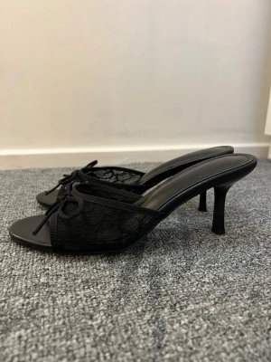 Svarta spetsiga pumps med rosett - Säljer ett par eleganta svarta pumps med spetsdetaljer och en fin rosett framtill. Skorna har öppen tå och smal klack, vilket ger en snygg och stilren look. Materialet är en kombination av mesh och syntet, perfekt för dig som gillar trendiga skor.
