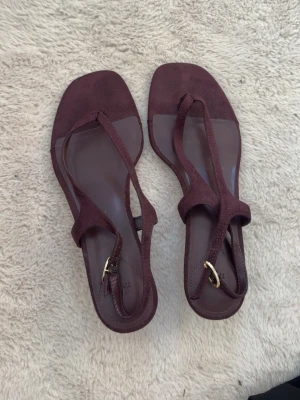 Vinröda sandaler med T-rem - Snygga vinröda sandaler med T-rem och justerbart spänne i guld. Sandalerna har en fyrkantig tå och är gjorda i mockaimitation. Perfekta för dig som vill ha en stilren och trendig look till sommaren. Endast använda en gång syns endast under skon