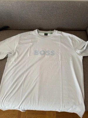 Grå BOSS t-shirt med broderad logga - Säljer en gråvit t-shirt från BOSS med stor broderad logga i ljusblått och vitt på bröstet. T-shirten har rund halsringning och är tillverkad med 100% bomull. Perfekt för en clean och stilren look.