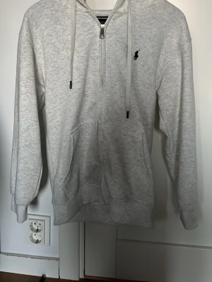  BYTE MOT RALPH LAUREN/ KÖPA   - Ljusgrå hoodie med dragkedja och huva från Polo Ralph Lauren. Klassisk design med känguruficka framtill, justerbara snören i huvan och broderad logga på bröstet. Perfekt för en avslappnad och stilren look.