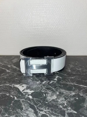 Vitt skärp med silverfärgat H-spänne - Snyggt vitt bälte med ett stort, silverfärgat H-format spänne. Bältet har en stilren design och är gjort i skinn med svart insida. Perfekt accessoar för att lyfta din outfit. **RESERVERAD**