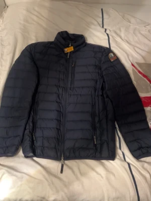Mörkblå dunjacka med patch - Snygg mörkblå dunjacka med quiltad design och hög krage. Jackan har dragkedja framtill, två sidofickor med dragkedja, två inne fickor och en bröstficka. Detaljerad patch på ena ärmen och gul rem vid kragen. Perfekt för kalla vinterdagar.