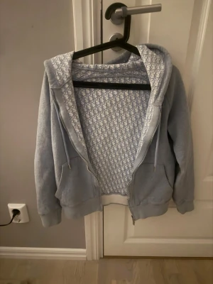 Ljusblå hoodie med dragkedja - Säljer en ljusblå hoodie med dragkedja och huva. Insidan har ett vitt och blått mönster som ger en unik touch. Hoodien har fickor framtill och justerbara snören i huvan. Perfekt för dig som gillar en avslappnad och stilren look.