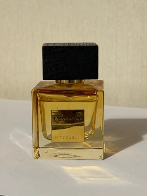 L'Essence parfym från Rituals - Elegant parfymflaska från Rituals, L'Essence. Flaskan är fyrkantig i genomskinligt glas med gyllene vätska och har en stor, svart träkork. Designen är stilren och lyxig, perfekt som en snygg detalj på sminkbordet.
