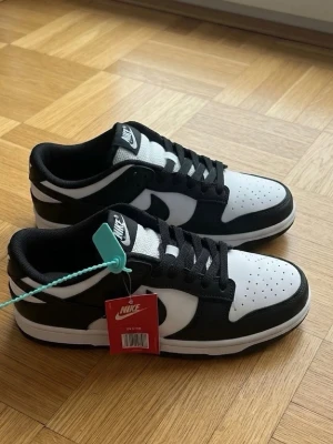 Ny Nike Dunk Low svart/vit sneakers - Nike Dunk Low sneakers i svart och vitt läder med klassisk swoosh på sidan. Skorna har perforerad tåbox, svart yttersula och vita detaljer. Låga och stilrena med svart snörning och vadderad plös. Perfekta för dig som gillar streetwear och enkel design.