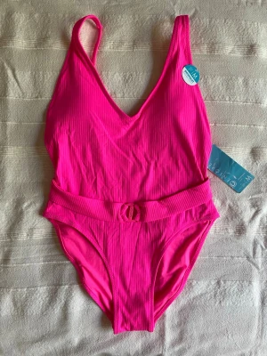 Rosa baddräkt med bälte och ribbad struktur, stl M - Snygg rosa baddräkt med ribbad struktur och ett matchande bälte i midjan. Djup v-ringning fram och smala axelband ger en trendig look. Materialet ser ut att vara stretchigt och bekvämt, perfekt för strand eller pool. Baddräkten har en modern och ungdomlig stil.