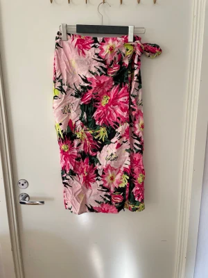 Blommig strandkjol i rosa och grönt, stl M oanvänt  - En lång strandkjol med knytning i sidan och färgstarkt blommönster i rosa, grönt, svart och gult på ljusrosa botten. Kjolen är luftig och lätt, perfekt att dra över bikinin. Materialet känns mjukt och följsamt, vilket gör den enkel att bära på stranden.