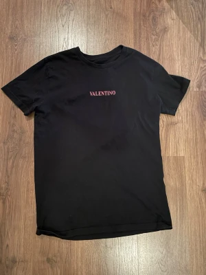 Svart t-shirt från Valentino - Snygg svart t-shirt från Valentino med diskret rosa logga på bröstet. Klassisk passform och rund halsringning. Tillverkad i mjuk bomull som känns skön mot huden. Perfekt till jeans eller annat för en clean och stilren look. Saknar storleksangivelse men passar som S/M
