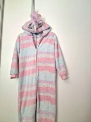 Randig enhörningsonesie med huva - Supermysig onesie i pastellrosa och ljusblått med fluffigt material. Dragkedja framtill och huva med enhörningshorn, öron, broderade ögon och rosa man. Perfekt för chillkvällar eller när du vill vara extra cozy.