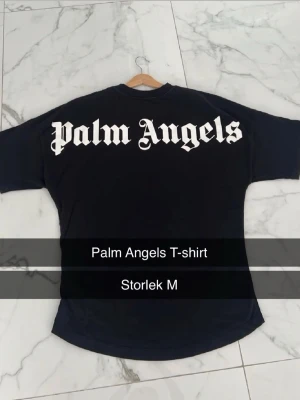 Svart Palm Angels T-shirt - Snygg svart T-shirt från Palm Angels med stort vitt logotryck i gotisk stil över ryggen och mindre logga i nacken. Oversized passform och rund hals. Perfekt för en streetwear-look.