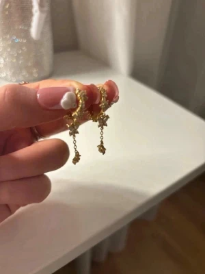 Guldiga örhängen med hängande stjärnor - Säljer ett par guldiga örhängen med små gnistrande stenar och hängande stjärndetaljer. Örhängena har en tunn kedja och ger en elegant look med sina fina detaljer. Perfekta för dig som gillar unika accessoarer med lite extra bling.