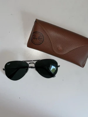 Ray-Ban Aviator solglasögon svart - Klassiska Ray-Ban Aviator solglasögon med svarta glas och tunn metallbåge i silver. Säljs med originalbrunt fodral. Perfekt accessoar för en cool och tidlös stil.
