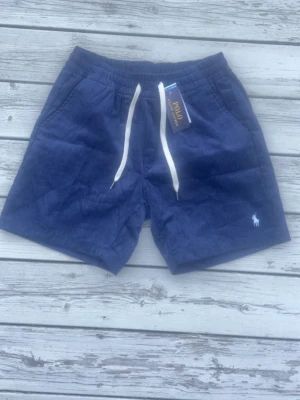 Marinblå manchester shorts Polo Ralph Lauren - Säljer ett par marinblå manchester shorts från Polo Ralph Lauren med vit snörning i midjan och klassisk logga broderad på benet. Shortsen har sidofickor och avslappnad passform, perfekta för sommardagar.