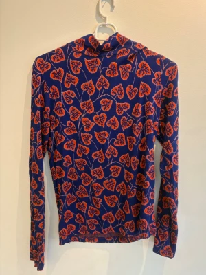 Blommig blå och röd polotopp - Snygg långärmad polotopp i blått med rött blommönster. Toppen har en tight passform och hög krage. Från Rodebjer.