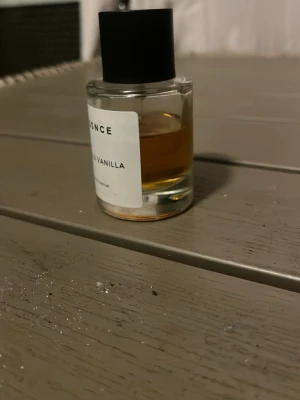 ESSNCE Absolu Vanilla parfym - En stilren parfymflaska från ESSNCE med doften Absolu Vanilla. Flaskan är cylindrisk i genomskinligt glas med svart lock och vit etikett med svart text. Innehållet har en varm, bärnstensfärgad ton. Perfekt för dig som gillar söta och mjuka dofter.