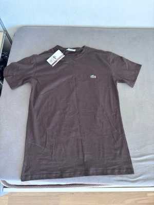 Brun t-shirt från Lacoste - Snygg brun t-shirt från Lacoste med klassisk krokodillogo på bröstet. T-shirten har rund hals och korta ärmar, perfekt för en clean och enkel stil. Tillverkad i mjuk bomull som känns skön mot huden.