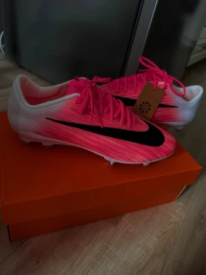 Nike rosa och vita fotbollsskor - Säljer ett par snygga fotbollsskor från Nike i en grym rosa och vit färgkombination med svart swoosh på sidan. Skorna har snörning, låg profil och dobbar för spel på gräs. Sulan har även detaljer i neonlime. Perfekta för dig som vill sticka ut på planen!