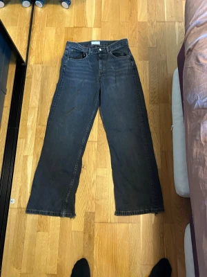 Svarta loose/bootcut jeans med råa kanter - Säljer ett par svarta jeans med rak passform och råa, fransiga benslut. Jeansen har klassisk femficksdesign och hög midja. Perfekta för en avslappnad och trendig look. Materialet är denim i bomull. Storlek 32/34