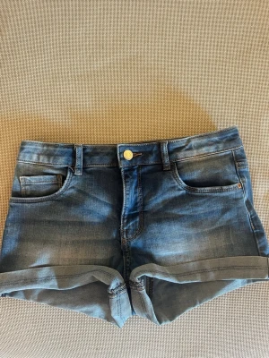 Lågmidjade blå jeansshorts med uppvik - Snygga blå jeansshorts med klassisk femficksdesign och uppvikta benslut. Shortsen är lågmidjade och har normal passform och stängs med knapp och dragkedja. Perfekta för varma dagar och enkla att matcha med olika toppar.