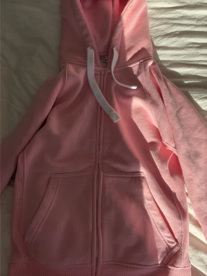 Rosa hoodie med dragkedja Russedress - Säljer en ljusrosa hoodie från Russedress med dragkedja framtill, vita snören i huvan och två stora fickor. Tillverkad i mjukt bomullsmaterial och har ribbade muddar vid ärmslut och nederkant. Perfekt för en chill och avslappnad stil.