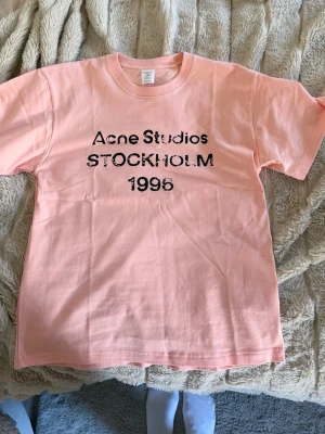 Ljusrosa Acne Studios t-shirt - Säljer en ljusrosa t-shirt från Acne Studios med svart tryck på bröstet där det står 'Acne Studios STOCKHOLM 1996'. Klassisk passform och rund halsringning. Perfekt för dig som gillar stilrena plagg med en twist.