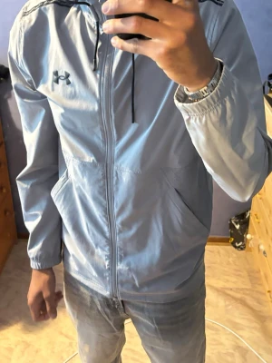 Ljusblå vindjacka från Under Armour - Snygg ljusblå vindjacka från Under Armour med dragkedja framtill, huva med dragsko och två sidofickor. Jackan har en sportig look med svart logga på bröstet och elastiska muddar vid ärmsluten. Perfekt för blåsiga dagar.