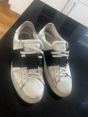 Vita sneakers med svart band Valentino - Snygga vita sneakers från Valentino med ett brett svart band över mitten och vita skosnören. Klassisk låg modell med rund tå och diskret logga på plösen. Perfekta för dig som gillar stilrena och trendiga skor.