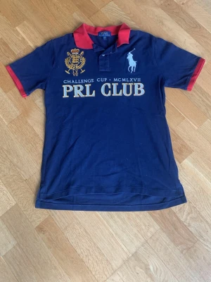 Mörkblå PRL Club pikétröja - Snygg mörkblå pikétröja med röd krage och röda ärmslut. Framtill finns broderade detaljer och texten 'PRL CLUB', samt en stor siffra 3 på ryggen. Klassisk polodesign med korta ärmar och knappslå. Perfekt för en sportig och avslappnad stil.