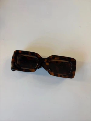 Rektangulära solglasögon leopardmönster - Snygga solglasögon med rektangulära bågar i leopardmönster. Glasen är mörka och bågarna har en cool, chunky design som ger en trendig vibe. Perfekta för att lyfta din stil under soliga dagar.