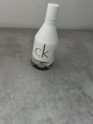 CK parfymflaska i vit och glas - Minimalistisk parfymflaska från CK med vit ovandel och genomskinlig glasbotten. Flaskan har en stilren design med tydlig CK-logga på framsidan. Perfekt för dig som gillar klassiska och moderna dofter.