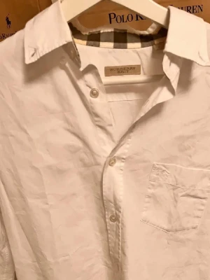 Vit skjorta från Burberry Brit - Klassisk vit skjorta från Burberry Brit med knappstängning och bröstficka. Insidan av kragen har det ikoniska rutiga Burberry-mönstret. Skjortan är tillverkad i bomull och har en tidlös, clean look litet hål i armvecket men går att fixa där av priset .