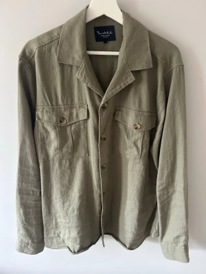 Olivgrön overshirt från Bondelid - Stilren olivgrön overshirt från Bondelid med ett mjukt linnematerial. Skjortan har två bröstfickor med knappar, klassisk krage och knäppning framtill. Perfekt lager på lager-plagg med avslappnad passform och långa ärmar.