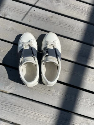 Valentino Garavani Open sneakers vit/mörkblå - Säljer ett par Valentino Garavani Open sneakers i vitt skinn med bred svart läderdetalj över snörningen. Klassisk rund tå och platt sula. Kommer inte  med originalpåse och box. Perfekta för dig som vill ha en clean men lyxig look. Jag kommer även att göra rent skorna mer senare.