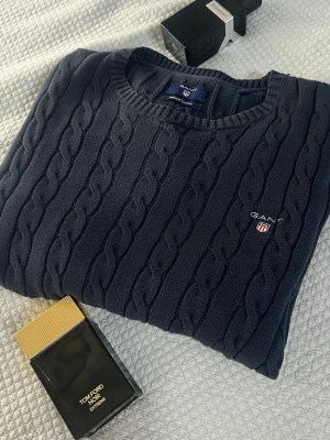 Mörkblå kabelstickad tröja från GANT - Klassisk mörkblå kabelstickad tröja från GANT med rund halsringning och diskret logga broderad på bröstet. Tröjan har långa ärmar och ribbade muddar vid ärmslut och nederkant. Perfekt för dig som gillar stilrena och tidlösa plagg.
