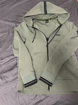 Ljusgrön hoodie med dragkedja från BOSS - Snygg ljusgrön hoodie från BOSS med dragkedja framtill, svarta detaljer och huva med snören. Ribbade muddar med svarta ränder och diskret BOSS-logga på bröstet. Perfekt för en avslappnad och trendig look.använd 2 gånger köpt på johnells för 2000 kan gå ner lite snabb affär