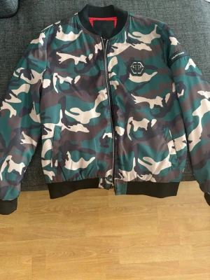 Camo bomberjacka från Philipp Plein - Snygg jacka i camomönster från Philipp Plein med svarta muddar och dragkedja framtill. Jackan har logga på bröstet och stor röd 'PLEIN' text på ryggen. Tillverkad i ett glansigt material som ger en cool streetwear.                                                                     