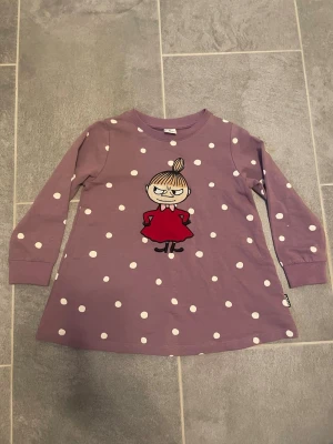 Lila klänning med Lilla My och prickar - Söt lila klänning med vita prickar och tryck av Lilla My på bröstet. Klänningen har lång ärm och är gjord i mjuk bomull. Perfekt för dig som gillar Mumin och vill ha något bekvämt och lekfullt i garderoben.