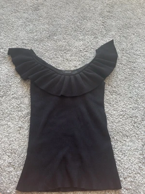 Svart topp med volang från Vero Moda - Trendig svart topp från Vero Moda med bred volang runt den djupa ringningen. Toppen är ribbad och har ett mjukt material. Vid kanten där uppe  har den gummi så att den inte ska glida ner 