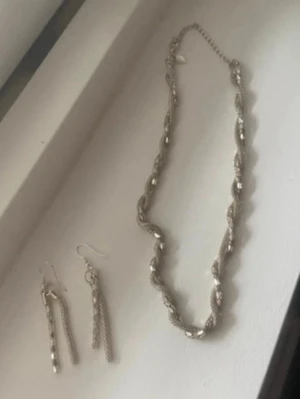 Silvrigt halsband och örhängen set - Säljer ett matchande set med ett silvrigt halsband och ett par örhängen. Halsbandet har en tvinnad kedja och örhängena har hängande kedjor i olika längder. Perfekt för dig som gillar stilrena accessoarer med lite edge.