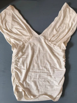 Gina tricot vit omlottopp med v-ringning - Säljer en vit omlottopp med djup v-ringning och rynkade sidor. Toppen har korta, vida ärmar och är tillverkad i mjukt bomullsmaterial. Perfekt för dig som gillar en feminin och stilren look.