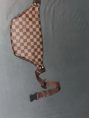 Brun rutig magväska - Snygg magväska i brunt och beige rutigt mönster. Väskan har justerbart brunt band med spänne och flera dragkedjefack framtill. Perfekt för att bära det viktigaste och ger en trendig streetstyle-look.